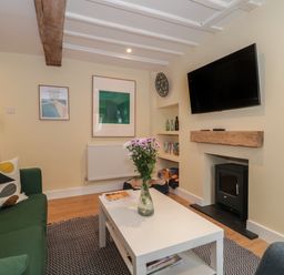 A living room with a green sofa a white coffee table with flowers a wall-mounted television and a small stove at Holly Cottage in West Coker