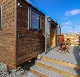 A wooden cabin with two windows and an open door leading to a deck with a wooden chair and table at Claerwen Nant-glas near Rhayader
