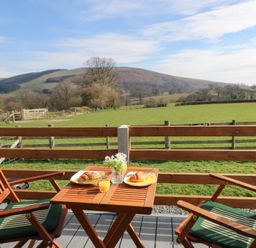 A wooden table with two croissants glasses of orange juice and a small flower vase on a patio with two wooden chairs overlooking fields and hills at Claerwen Nant-glas near Rhayader
