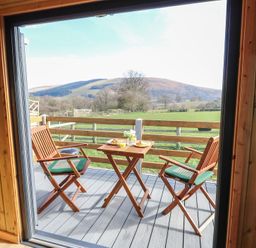 A balcony with two wooden chairs and a small table set with food and drinks overlooking fields and hills at Claerwen Nant-glas near Rhayader