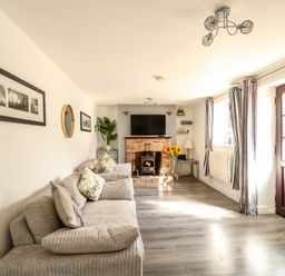 A living room with a sofa, pillows, a brick fireplace with a wood stove, a TV above the fireplace, a door, windows with curtains, a floor lamp, and framed pictures on the wall at Chalk Cottage in Feltwell