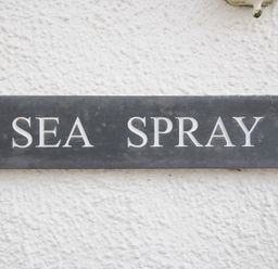 A black rectangular sign with white letters reading SEA SPRAY mounted on a white textured wall at Sea Spray in Port Isaac