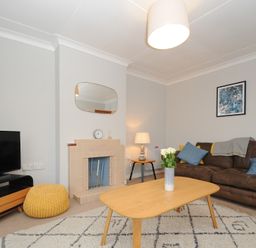 A living room with a brown sofa with blue and yellow cushions a light wooden coffee table with flowers a TV on a stand a fireplace and a lamp at Sea Spray Port Isaac