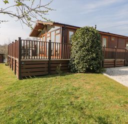 A wooden cabin with a deck and railing surrounded by grass and shrubs at Meadow Lodge in Newton Ferrers