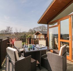 An outdoor deck with a table and four chairs next to a wooden building with glass doors at Meadow Lodge in Newton Ferrers