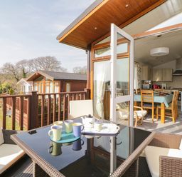 A patio with a black glass table and four chairs overlooking wooden cabins and a dining area inside with a table and chairs at Meadow Lodge in Newton Ferrers