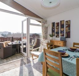 A dining area with wooden chairs and a table set with plates and glasses next to a door opening to an outdoor seating area at Meadow Lodge in Newton Ferrers