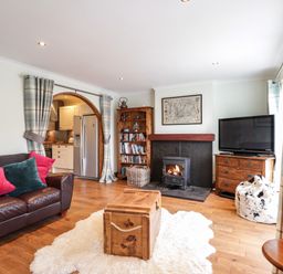 A living room with a leather sofa and colorful pillows a wooden chest on a rug a stove fireplace a bookshelf a tv on a dresser and curtains at The Hideout in Portree
