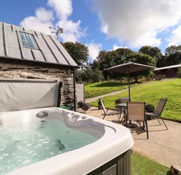 An outdoor patio with a hot tub a table with four chairs and an umbrella next to a stone building with a metal roof at Y Felin at Coed Cadw in Felindre Farchog near Newport Pembrokeshire