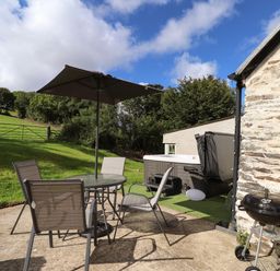 An outdoor patio with a table and four chairs under an umbrella a barbecue grill and a hot tub next to a stone building at Y Felin at Coed Cadw in Felindre Farchog near Newport Pembrokeshire