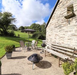 An outdoor patio with a metal table and chairs with an umbrella a barbecue grill several potted plants and a wooden bench by a stone wall at Y Felin at Coed Cadw in Felindre Farchog near Newport Pembrokeshire