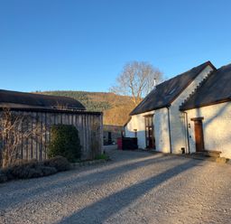 An exterior view of a white stone building with a gravel driveway and a wooden barn with hills in the background at Maes Y Berllan Barn in Gilwern