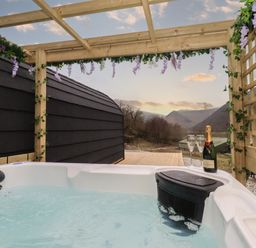 An outdoor hot tub with a bottle of champagne and two glasses under a wooden pergola with flowers at Jenny - Crossgate Luxury Glamping in Hartsop near Glenridding
