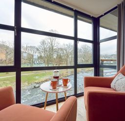 A seating area with two orange chairs and a small round table with a coffee press and two mugs by large windows overlooking a river at Riverside Park 6 in Keswick