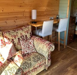 A wooden interior room with a patterned sofa a tall wooden table with two chairs and a bed visible through a door at Lime Kiln in Blagdon