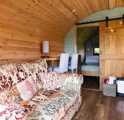 An interior view of a wooden room with a patterned sofa, a table with two white chairs, and a sliding wooden door leading to a bedroom at Lime Kiln in Blagdon