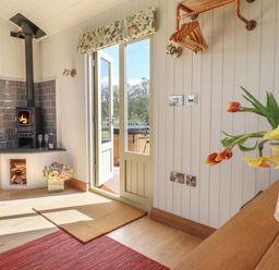 An interior with a wood stove, open door leading outside, wall-mounted clothes hangers, and flowers on a wooden surface at Balwen Shepherd's Hut in Berriew