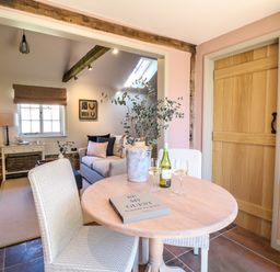 A dining area with a round wooden table, two chairs, a book, a vase with branches, two wine glasses, and a bottle of wine next to a wooden door and a living room with a couch and a window at The Forge in Old Newton near Stowmarket