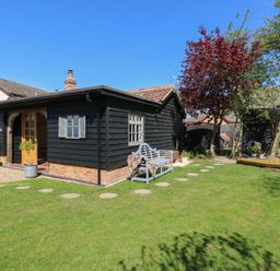 A black wooden garden shed with small windows and a door surrounded by grass and trees at The Forge in Old Newton near Stowmarket