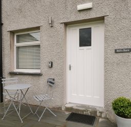 A white door and window with blinds outside with a small table and two chairs on a stone patio and a potted plant at Bill's Barn in Oxen Park near Ulverston