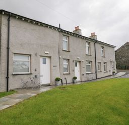 A row of connected houses with white doors and windows and small outdoor tables and chairs on a paved path with a grassy area at Bill's Barn Oxen Park near Ulverston