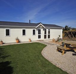 An exterior view of a single-story building with a grass lawn a gravel path and a wooden picnic table in the yard at Arwelfa Lodge in Abersoch
