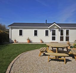 A garden with a wooden picnic table on a gravel area and a white house with multiple windows at Arwelfa Lodge in Abersoch