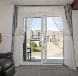 A sliding glass door with sheer curtains overlooking a street with houses at Bywyd Da (232 Cae Du) in Abersoch