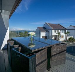 An outdoor balcony with a glass table and wicker chairs overlooking houses at Cromlin Glas in Abersoch