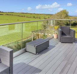 An outdoor deck with two wicker chairs a wicker footrest and glass railing overlooking grassy fields at Eleri in Abersoch