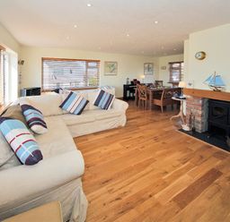 A living room with a beige sectional sofa and striped cushions near windows a dining table and chairs a fireplace with a wood stove and wooden floor at Eleri in Abersoch