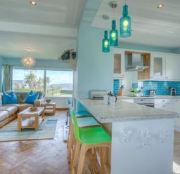 A living room with a brown sofa and blue pillows next to a kitchen with a white counter and green and blue chairs at Gilfach in Abersoch