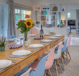 A dining room with a wooden table set with plates glasses cutlery and flowers surrounded by colorful chairs at The Coach House in Abersoch