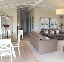 A dining area with a glass table and white chairs next to a living room with gray sofas and paintings on the wall at The Warren D26 in Abersoch