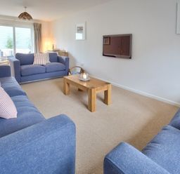 A living room with blue sofas a wooden coffee table a wall-mounted TV and sliding glass doors at Ty Coed in Abersoch