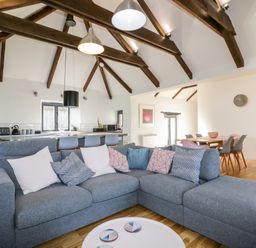 An open plan living area with a grey sectional sofa with cushions a round coffee table a dining table with chairs and a kitchen with bar stools and visible ceiling beams at Beacon Barn in St Agnes
