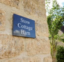 A sign reading Stow Cottage Barn on a stone wall with trees and a fence in the background at Stow Cottage Barn in Churchill Oxfordshire