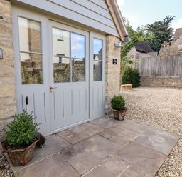 A stone house entrance with a grey door flanked by two potted plants on a stone patio at Stow Cottage Barn in Churchill Oxfordshire