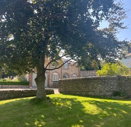 A garden with a large tree and a stone wall with a brick building in the background at The Coach House at Lackington Farm in Piddletrenthide