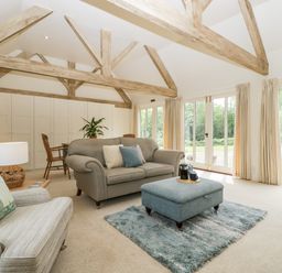 A living room with exposed wooden beams a sofa armchair ottoman and large windows at The Shippon in Honiton