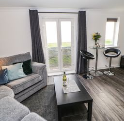 A living room with a grey sofa a black coffee table with a wine bottle and glass two black bar stools a round glass table with flowers and a kettle and a television at Sandy Corner in Blyth Northumberland