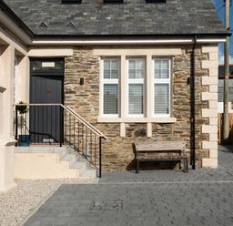 An exterior view of a stone house with three windows, a black door accessed by stairs, a bench, and a paved driveway at Church Rooms in Port Isaac