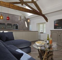 A living area with a blue sofa a glass coffee table with drinks and popcorn a TV on a gray cabinet and kitchen appliances at The Barn 20 At The Beach Torcross