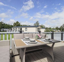 A patio deck with a wicker dining table set with plates bowls and flowers overlooking mobile homes at Yealands 27 South Lakeland Leisure Village near Carnforth