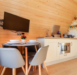 A small kitchen and dining area with a wall-mounted TV a round table set with bowls and plates and a kitchenette with a microwave and sink at Sea View in St Buryan