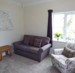 A living room with a purple sofa and plaid cushions a floral armchair a lamp and a window with purple curtains at Hafod Cottage in Cemaes Bay