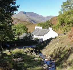 A white house near a small stream with trees and mountains in the background at Brookside in Grizebeck