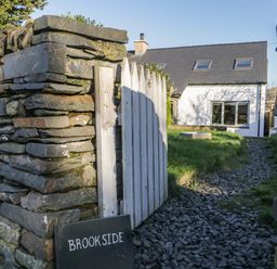 A stone wall with a white wooden gate and a sign reading Brookside with a path leading to a white house at Brookside in Grizebeck