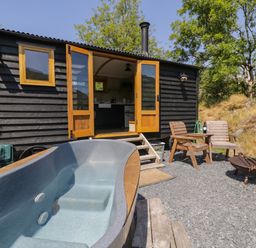 An outdoor area with a wooden hut opening to a kitchen an empty hot tub wooden chairs with a table holding a wine bottle and glass and a fire pit at Llethr Shepherds Hut in Llanbedr