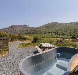 An outdoor area with a hot tub and wooden picnic table with mountains in the background at Llethr - Shepherds Hut in Llanbedr